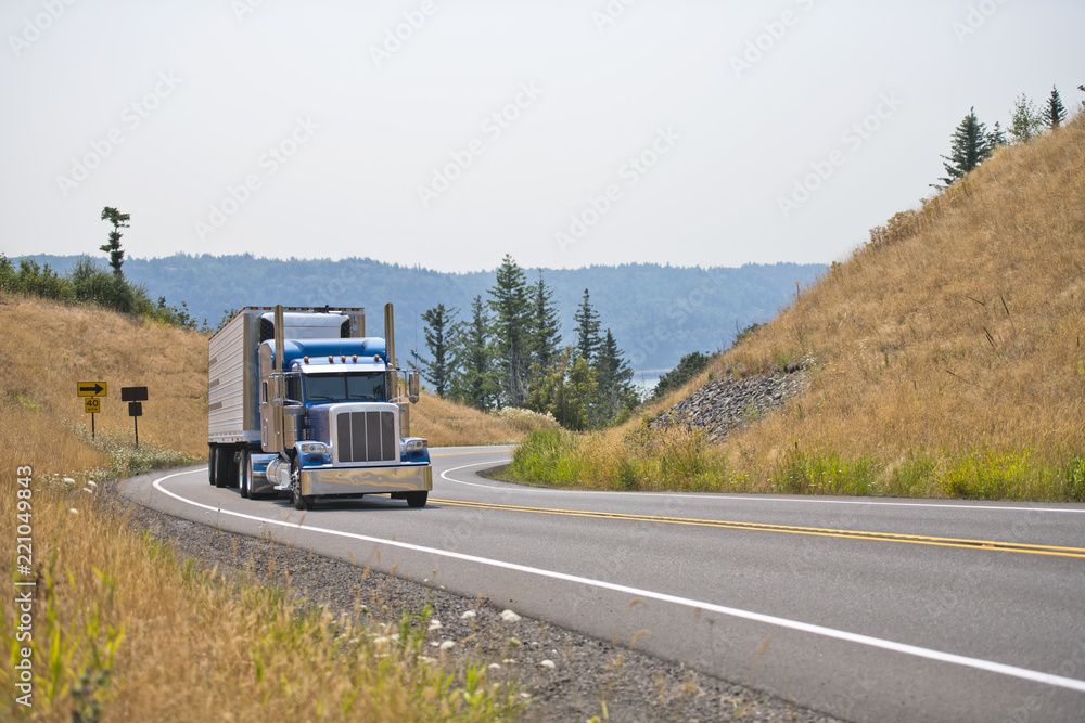 Blue big rig classic semi truck with refrigerated semi trailer turning on winding mountain highway