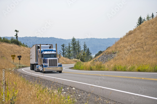 Blue big rig classic semi truck with refrigerated semi trailer turning on winding mountain highway