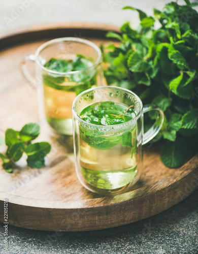 Hot herbal mint tea drink in glass mugs over wooden tray with fresh garden mint leaves, selective focus, close-up