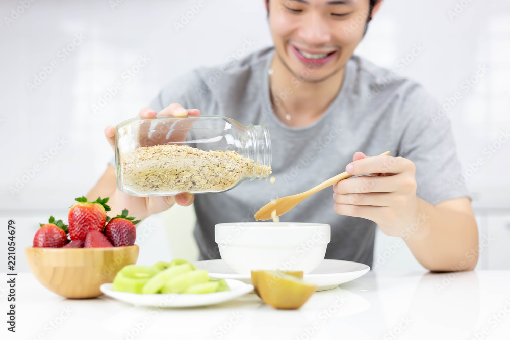 Attractive handsome young guy pouring cereal from glass bottle to bowl for making breakfast in the morning. Handsome man hold wooden spoon. Charming handsome guy eat with strawberry, kiwi fruit, milk