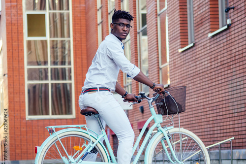 Fototapeta Naklejka Na Ścianę i Meble -  handsome African American man stylish well-dressed standing with a blue bicycle red brick building background . sports ecological transport concept