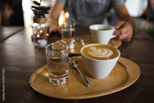 Cup of coffee cappuccino and a glass of water on a wooden tray. A man holding serving cup of coffee on the background