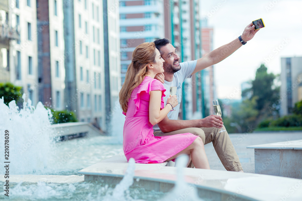 Our photo. Nice cheerful man taking a selfie while sitting together with his girlfriend