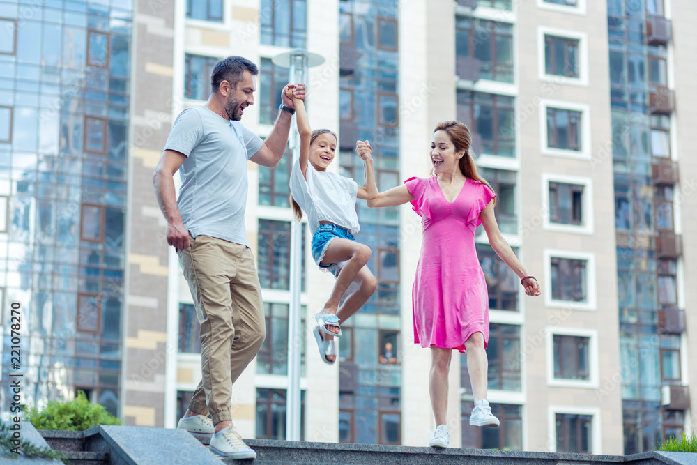 Playful child. Happy nice girl jumping while holding her parents hands