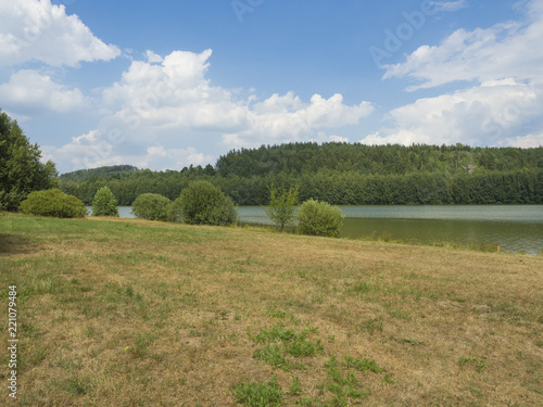 summer landscape with green lake or pond with spruce tree hill  sandstne rock and grass meadow trees  blue sky  white clouds