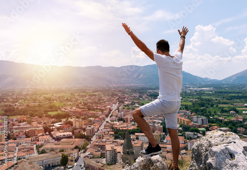 Man standing on the top of the mountain with his hands up looking at beautiful sunset and cityscape. Success concept