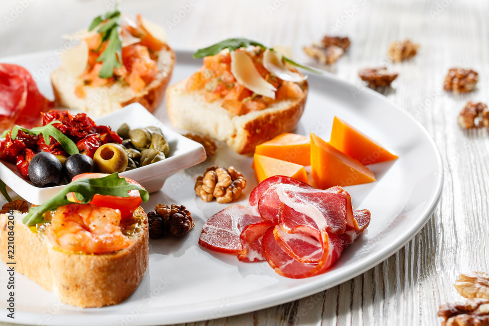 Plate with Italian appetizers. Bruschetta with a cherry tomatoes and shrimps. Parmesan cheese, prosciutto, green capers, olives, sun-dried tomatoes and walnuts. Plate with Italian appetizers. Bruschetta with a cherry tomatoes and shrimps. Parmesan cheese, prosciutto, green capers, olives, sun-dried tomatoes and walnuts.