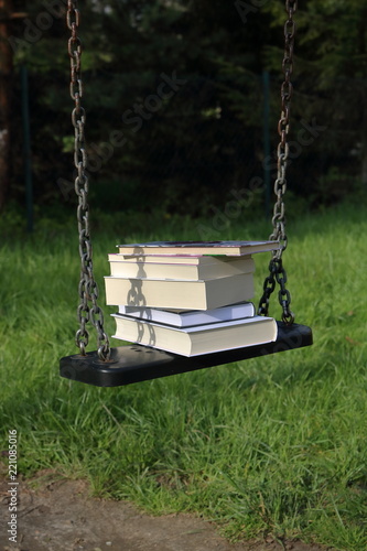 książki ułozone jedna na drugiej, kilka, leżą na dziecięcej huśtawce na łańcuchach, w ogrodzie