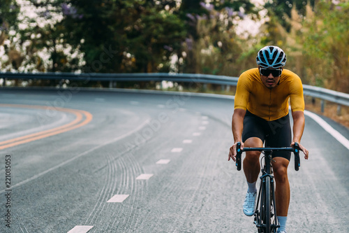 Fototapeta Naklejka Na Ścianę i Meble -  Focus on Asian man wearing a yellow cycling jersey, who's riding a road bike up high on hill in the morning. Under morning sunshine with determination on his face.