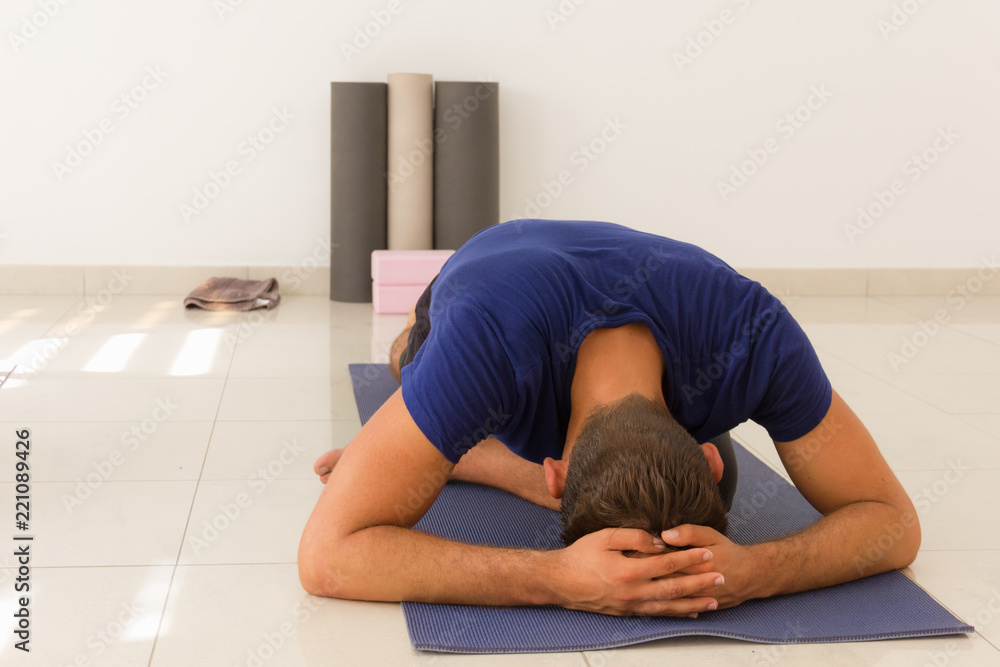 Male yogi in eka pada rajakapotasana on purple mat. Young man practices one legged king pigeon yoga pose indoors. Hands on head, relaxation, stretching, exercise, flexibility concepts