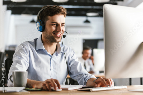 Photo of young worker man 20s wearing office clothes and headset, smiling and talking with clients in call center