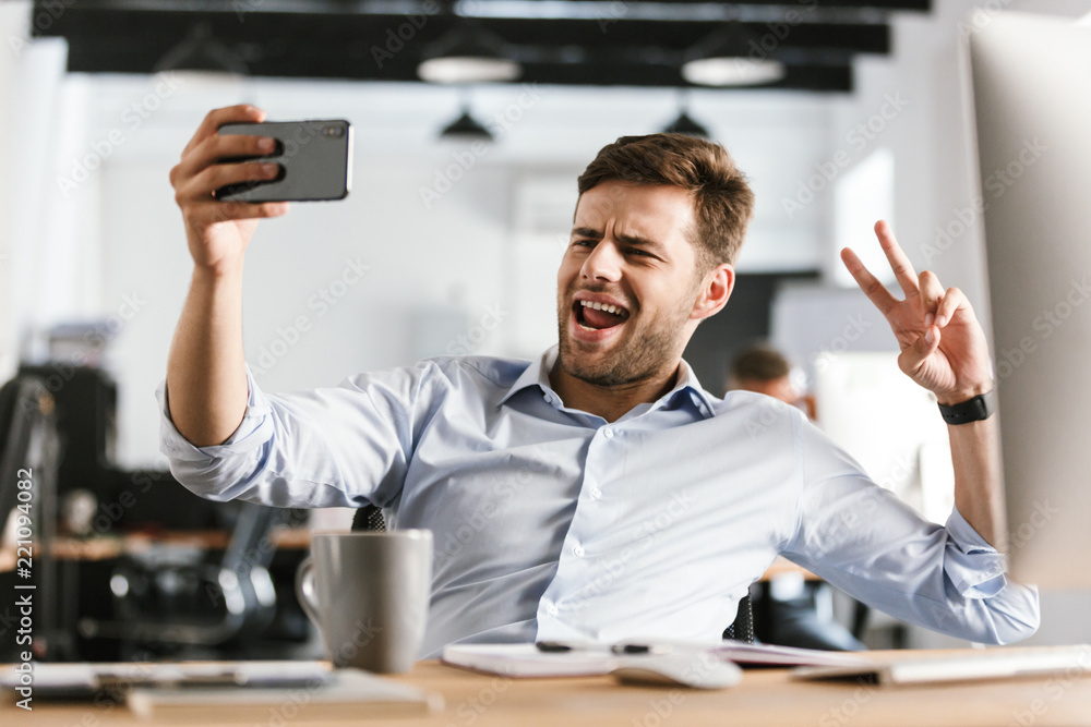 Fototapeta premium Playful business man making selfie on smartphone and showing peace