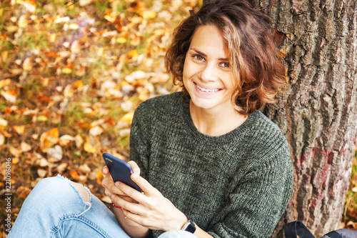 Beautiful young smiling  student girl woman with wavy hair sitting on grass and on fallen autumn leaves with smartphones in hands