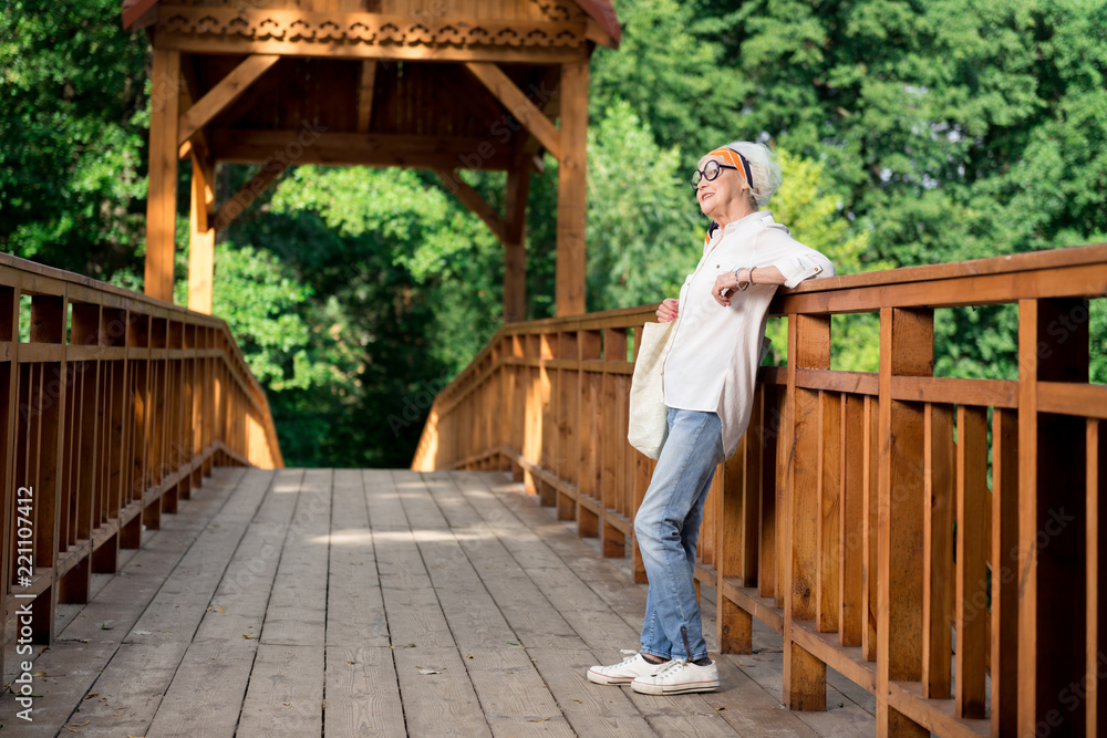 Jeans and sneakers. Stylish elderly woman wearing fashionable blue jeans and comfortable white sneakers walking in park