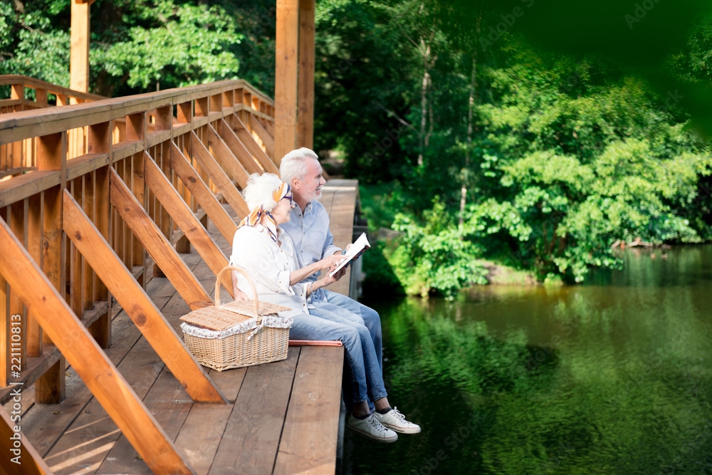 Bridge and hamper. Elderly wife and husband sitting on the wooden bridge near hamper while having traditional picnic Bridge and hamper. Elderly wife and husband sitting on the wooden bridge near hamper while having traditional picnic
