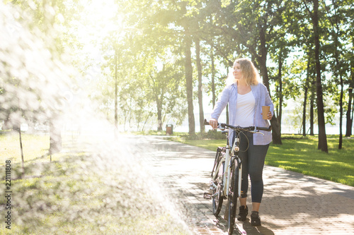 Young blonge woman walking her bicycle in the park and holding a cup of coffee