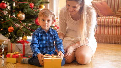 Cheerful toddler boy with smiling mother sitting under Christmas tree and looking on gifts and presents