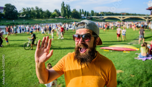 Happy to meet you. Urban event celebration. Hipster in cap happy to meet friend at event picnic fest or festival. Man bearded hipster in front of crowd people waving hand green riverside background