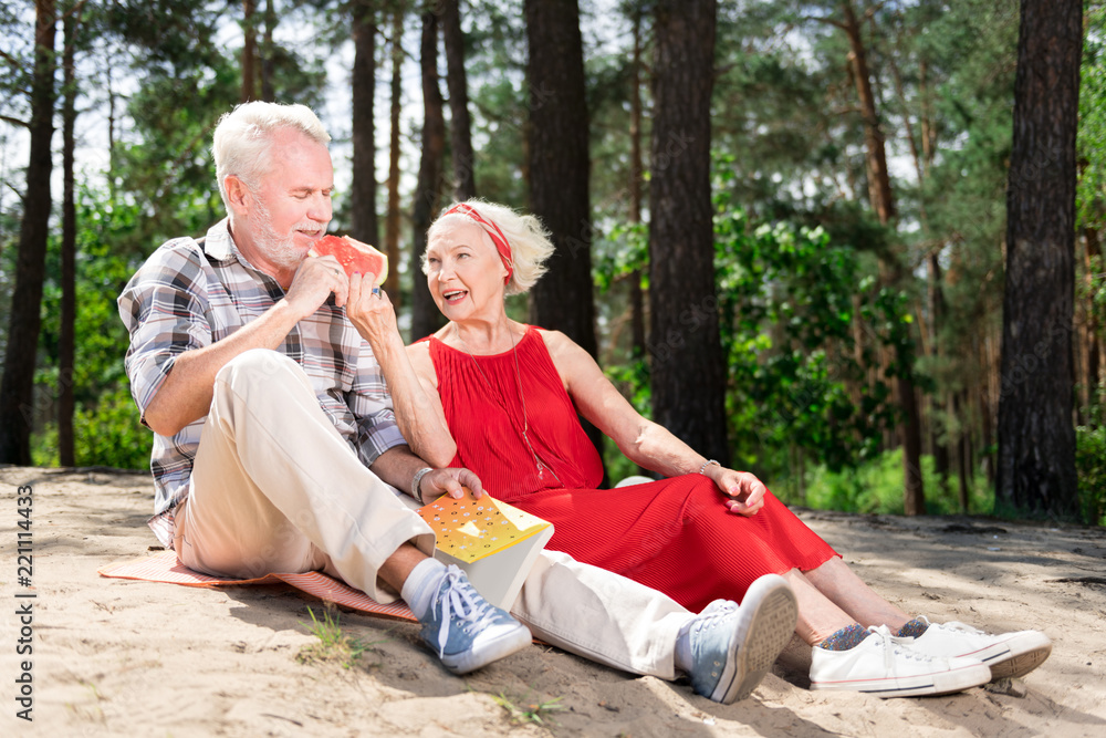 Cute pensioners. Cute beaming pensioners feeling rested while wearing sneakers eating water melon during picnic