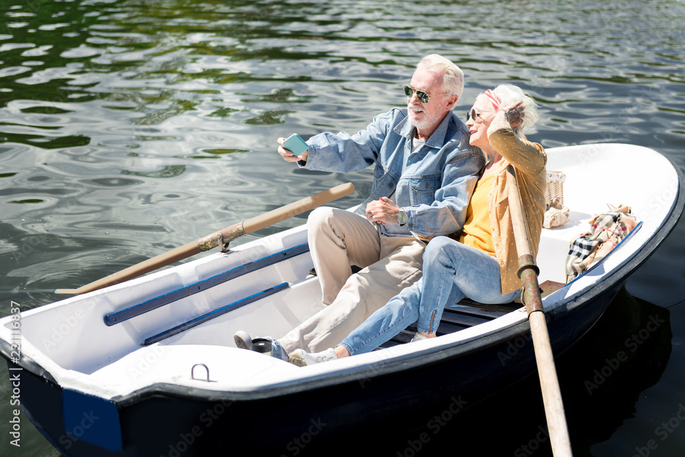Fototapeta premium Posing for photo. Beaming elderly lady posing for photo with her bearded stylish husband while sitting in boat