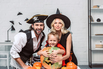 portrait of happy parents and son in halloween costumes at table with pumpkins at home