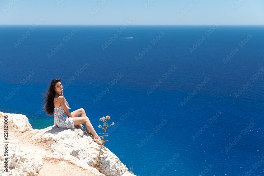 Brunette in a dress sits on the edge of the cliff overlooking the sea