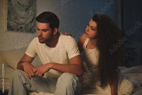 young woman looking at upset boyfriend in pajamas sitting on bed at night, relationship difficulties concept