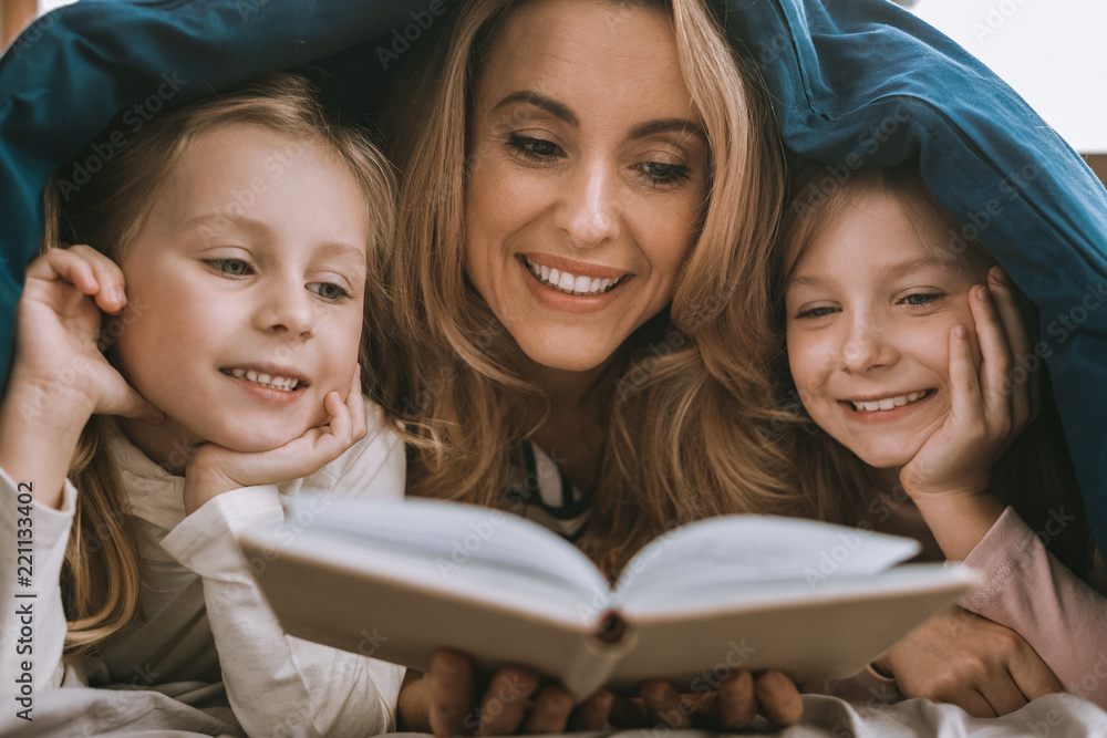 Time for fairytales. Delighted nice woman holding a book while resign fairytales to her daughters Time for fairytales. Delighted nice woman holding a book while resign fairytales to her daughters