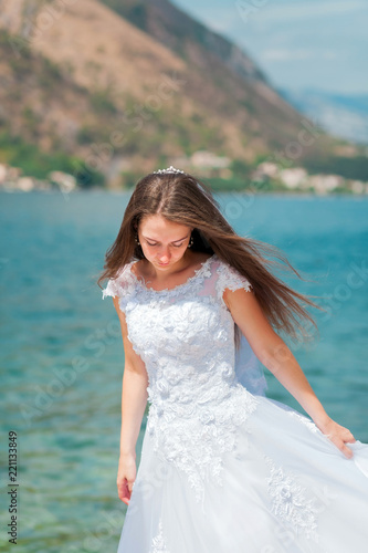 Portrait of a bride in a white wedding dress on a background of the sea and mountains. Girl brown-haired with long straight hair. Young beautiful woman looking down