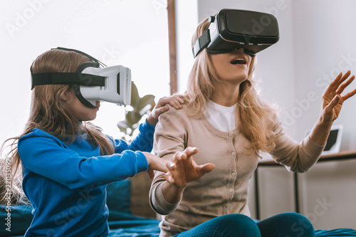 Technological developments. Nice young woman being in the virtual reality while sitting on the bed together with her daughter