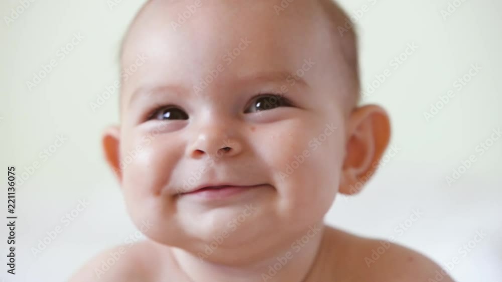 A cute little baby is looking into the camera and is happy on a white bed sheet smiles close up portrait A cute little baby is looking into the camera and is happy on a white bed sheet smiles close up portrait