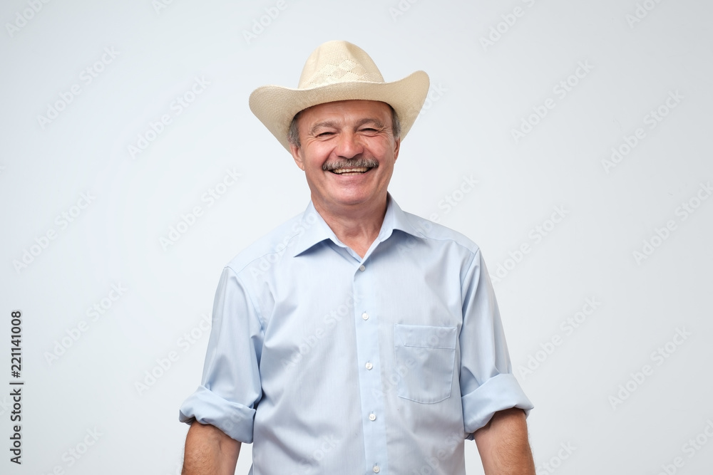 Cowboy style. Mature man adjusting his cowboy hat and laughing on joke while standing against grey background. Positive Cowboy style. Mature man adjusting his cowboy hat and laughing on joke while standing against grey background. Positive