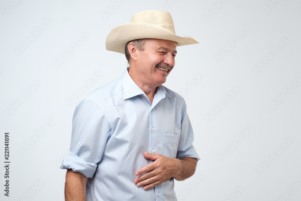 Cowboy style. Mature man adjusting his cowboy hat and laughing on joke while standing against grey background. Positive Cowboy style. Mature man adjusting his cowboy hat and laughing on joke while standing against grey background. Positive