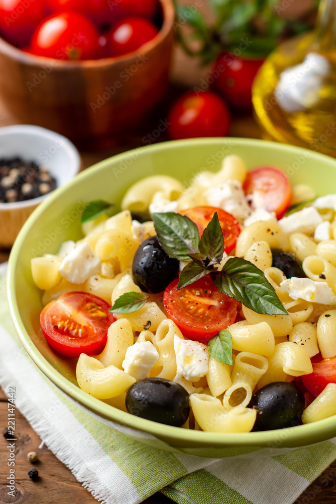 Pasta salad with cherry tomatoes, black olives, feta cheese and basil on wooden background