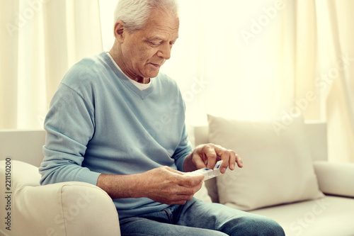 My prescription. Focused retired man sitting on a sofa and looking at a pill organizer in his hands while taking his medication.