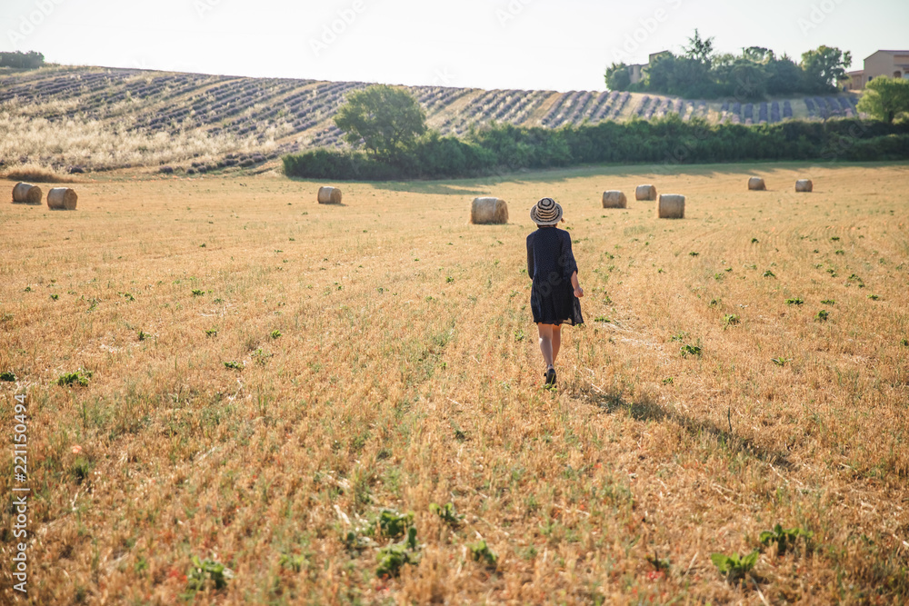 back view of girl in hat walking on agricultural field with hay bales, provence, france