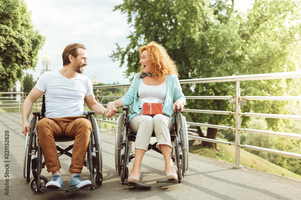 Excited news. Shocked woman looking at her husband with her mouth wide opened while both sitting in their wheelchairs outdoors. Excited news. Shocked woman looking at her husband with her mouth wide opened while both sitting in their wheelchairs outdoors.
