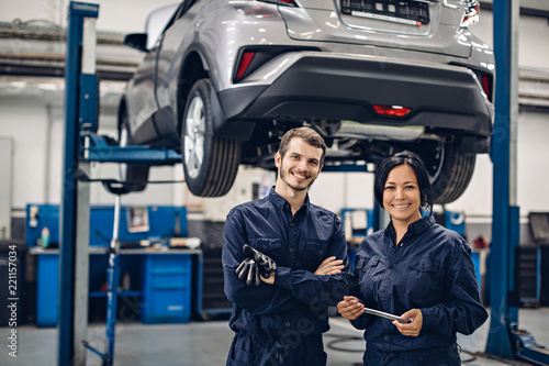 Auto car repair service center. Two happy mechanics - man and woman standing by the car