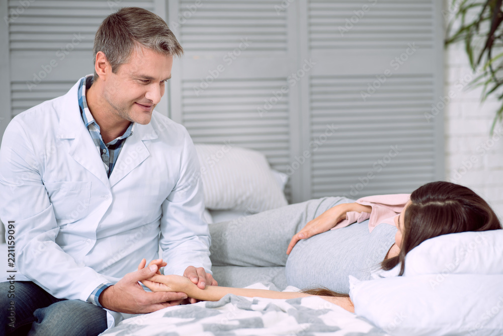Professional medicine. Positive nice professional doctor looking at his patient and checking the pulse while looking after her Professional medicine. Positive nice professional doctor looking at his patient and checking the pulse while looking after her