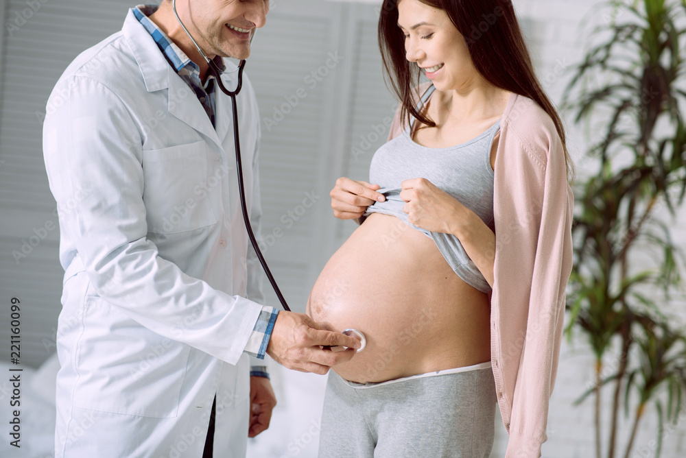 Medical equipment. Cheerful nice positive doctor holding a stethoscope and smiling while using it during medical checkup Medical equipment. Cheerful nice positive doctor holding a stethoscope and smiling while using it during medical checkup