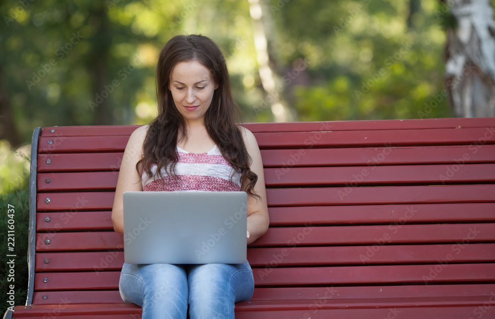 Obraz premium Beautiful caucasian woman is sitting at the bench in a city park with a laptop connected to wi-fi. Entrepreneur is working on a portable computer while sitting outdoors on a blurred urban background.