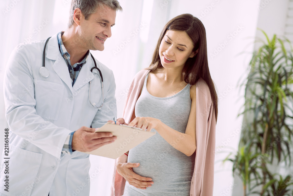 Pregnancy observation. Cheerful nice delighted woman holding her belly and pointing at the doctors notes while caring about her health Pregnancy observation. Cheerful nice delighted woman holding her belly and pointing at the doctors notes while caring about her health