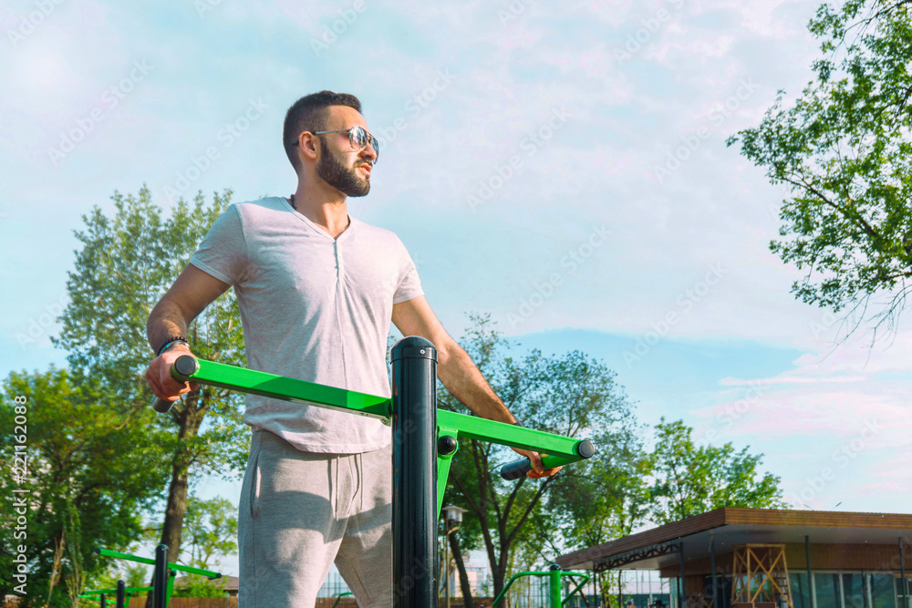 Fototapeta premium a young guy in good physical shape with a beard and in sunglasses trains on the simulators on the street in a city park on a hot sunny day in the summer