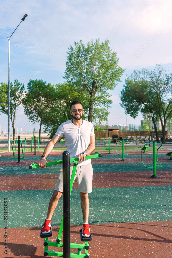 Obraz premium a young guy in good physical shape with a beard and in sunglasses trains on the simulators on the street in a city park on a hot sunny day in the summer