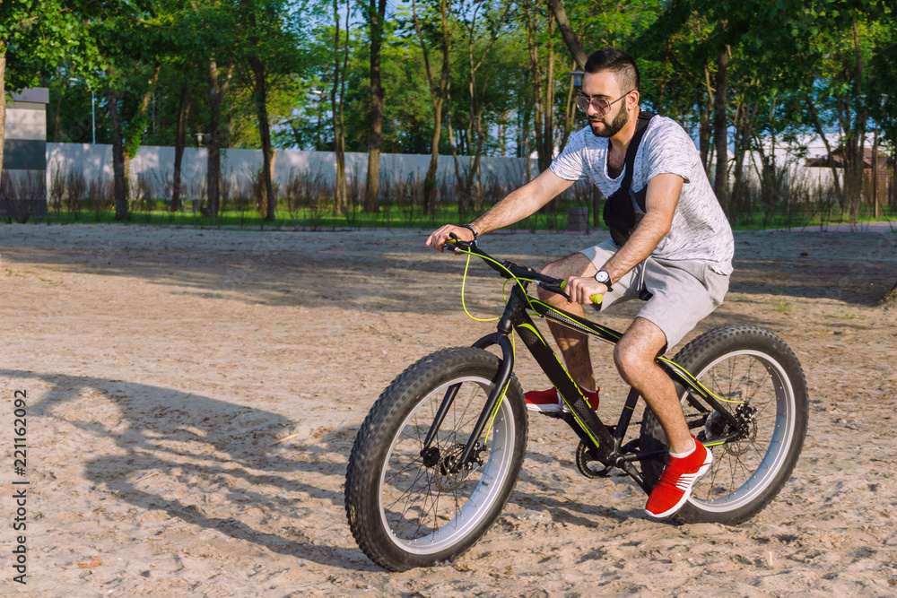 a young guy with a beard and wearing sunglasses on a big bike confidently skates through the city park on a hot, sunny day in the summer