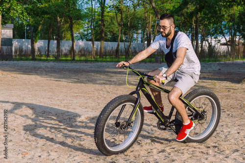 a young guy with a beard and wearing sunglasses on a big bike confidently skates through the city park on a hot, sunny day in the summer