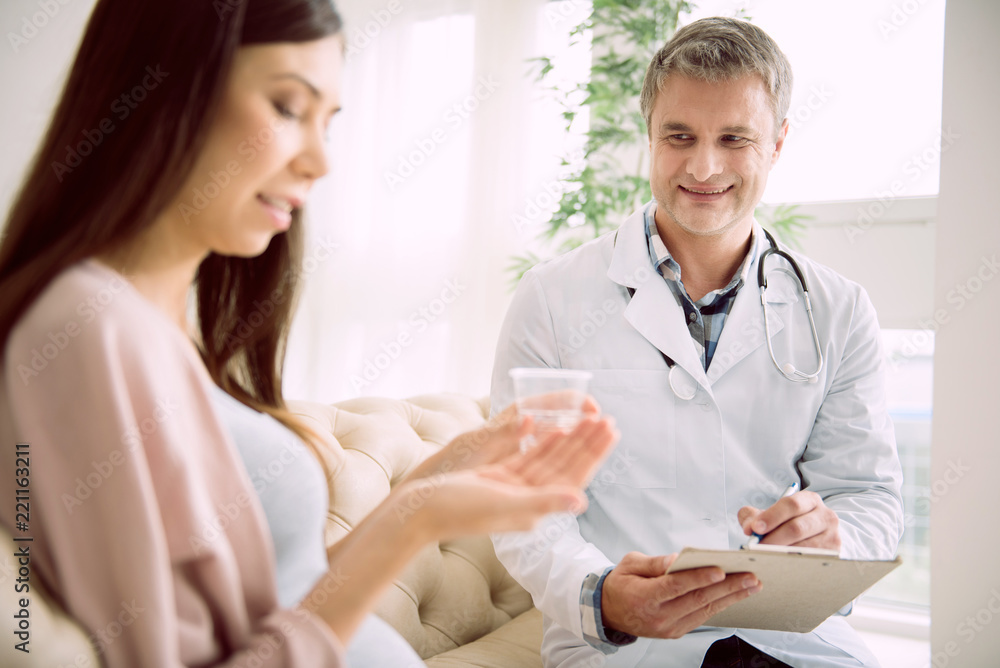 Professional therapist. Delighted nice skillful doctor smiling and observing his pregnant patient while taking notes Professional therapist. Delighted nice skillful doctor smiling and observing his pregnant patient while taking notes