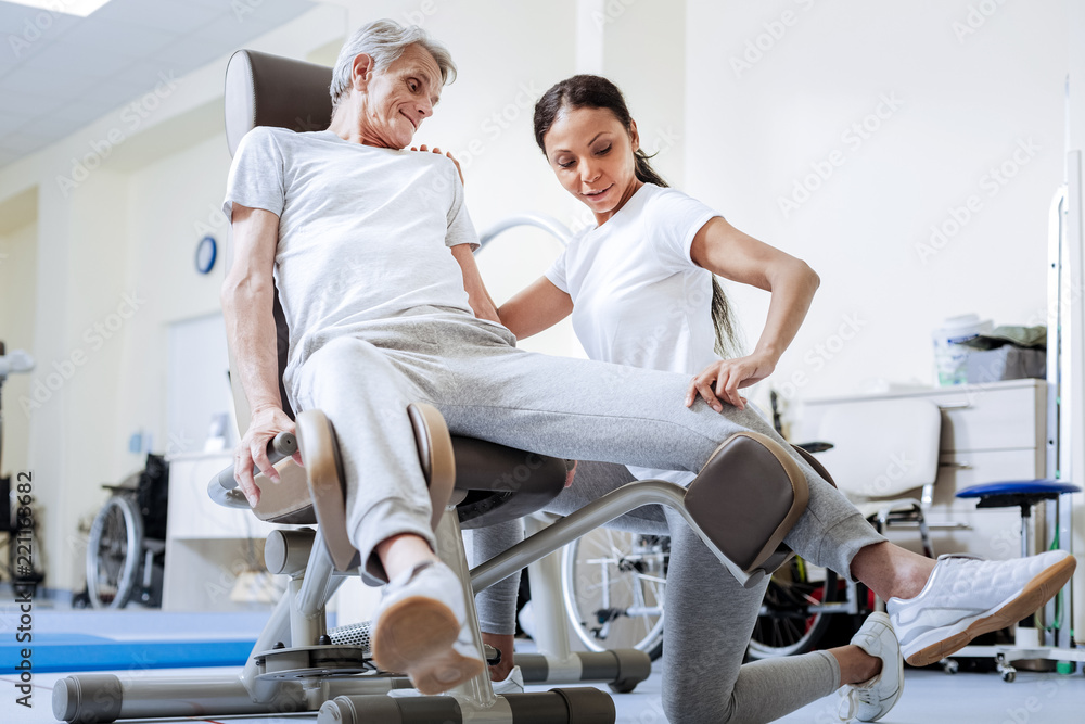 Obraz premium Professional help. Calm cheerful experienced doctor in a rehabilitation center touching the knee of her aged disabled patient while helping him to overcome his pain