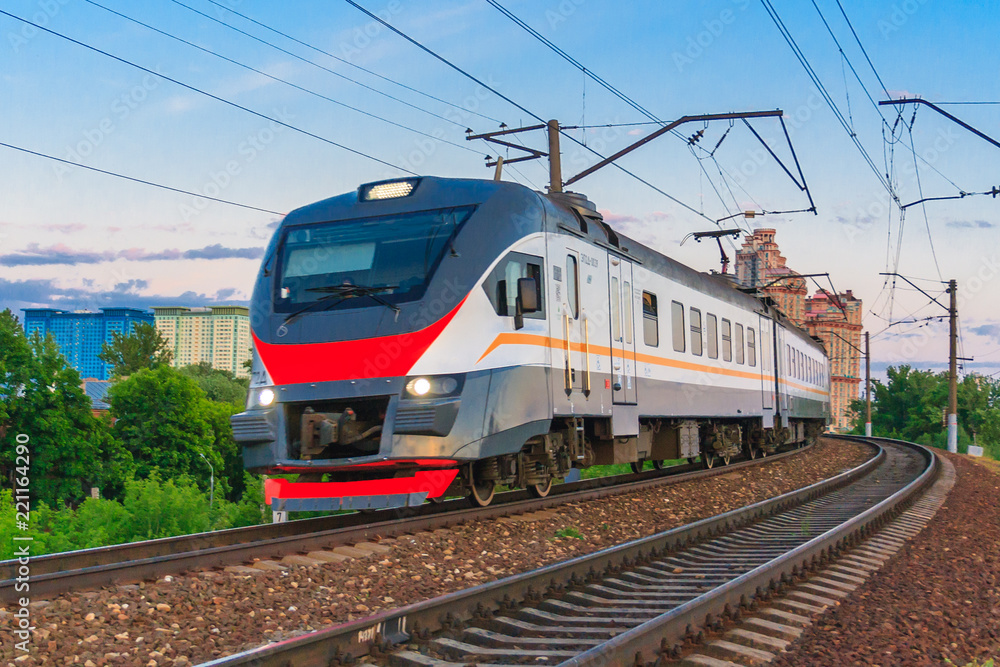 Passenger electric train with luminous headlights moves against the background of green trees and blue sky