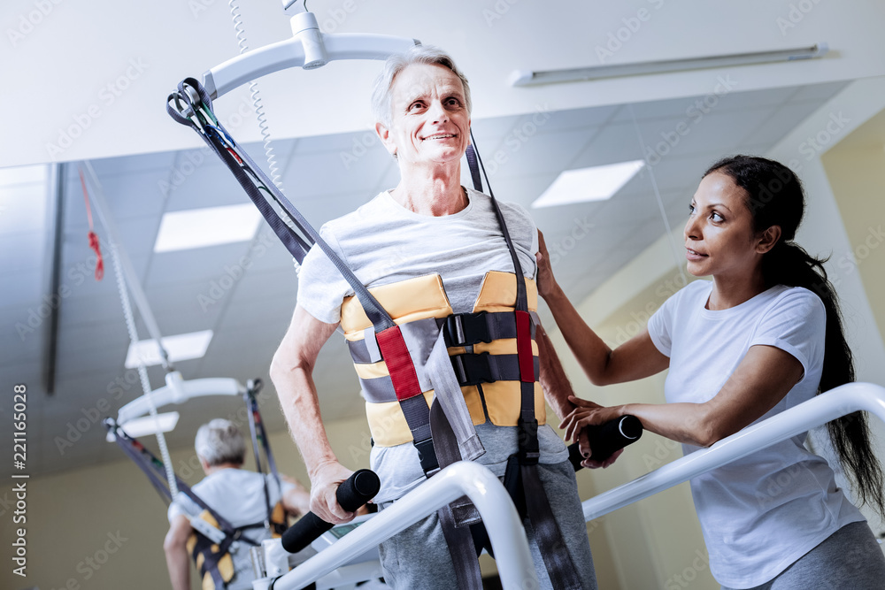 Feeling inspired. Positive smiling aged patient wearing a necessary body vest while going through the process of rehabilitation after a terrible injury with a kind attentive reliable medical worker Feeling inspired. Positive smiling aged patient wearing a necessary body vest while going through the process of rehabilitation after a terrible injury with a kind attentive reliable medical worker